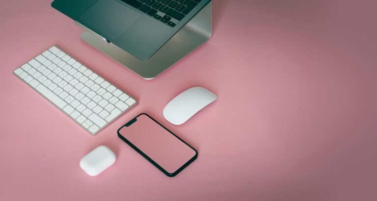 A minimalist desk setup featuring a laptop, keyboard, mouse, and smartphone on a pink background.
